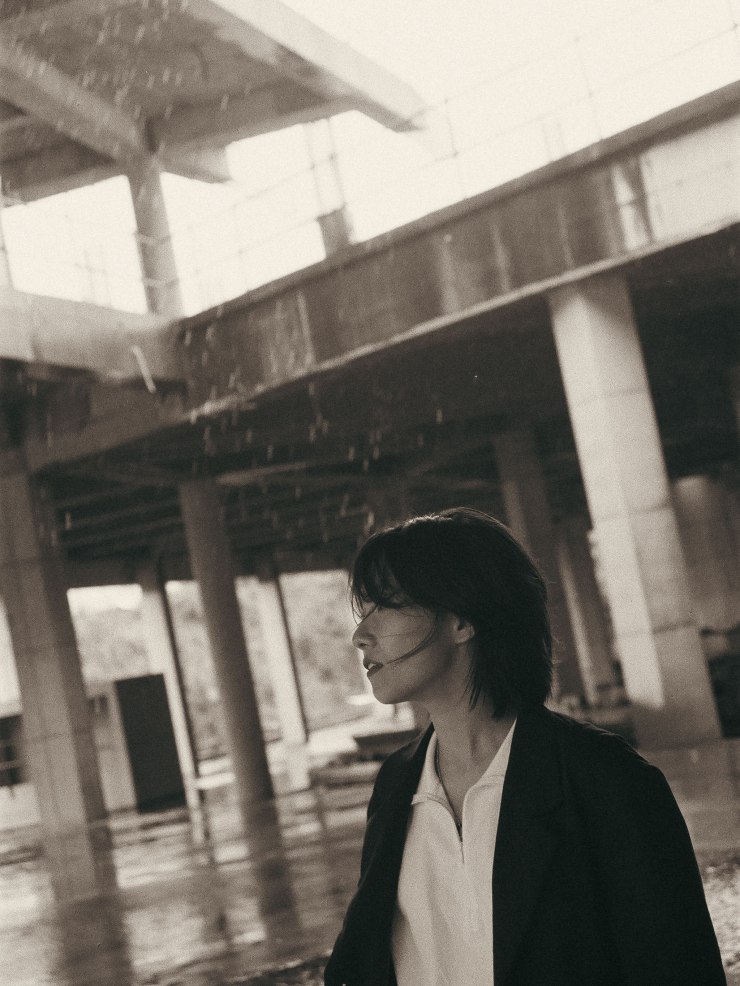Une femme se tient dans un espace industriel sous la pluie, avec des poutres et des colonnes en arrière-plan. Elle porte un haut blanc et un blazer noir, et ses cheveux sont légèrement mouillés, flottant au vent.