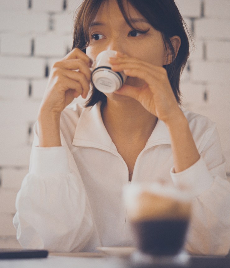 Une jeune femme boit dans une tasse en regardant pensivement, avec une ambiance intérieure douce et minimaliste en arrière-plan.
