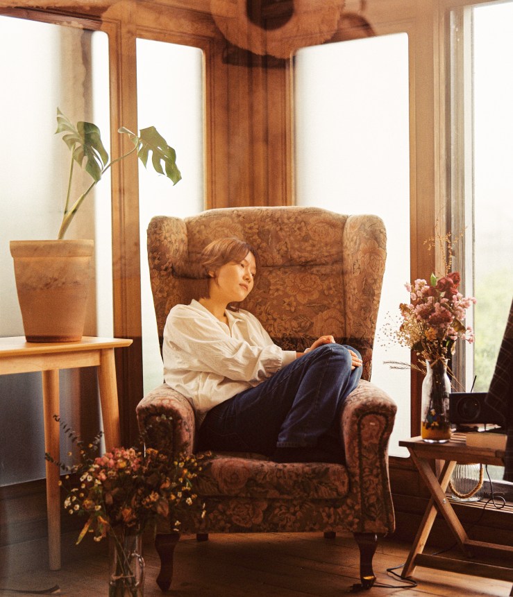 Une femme assise dans un fauteuil confortable, vêtue d'une chemise blanche et de jeans, regardant pensivement par une grande fenêtre. À côté d'elle, un vase avec des fleurs et une petite table.
