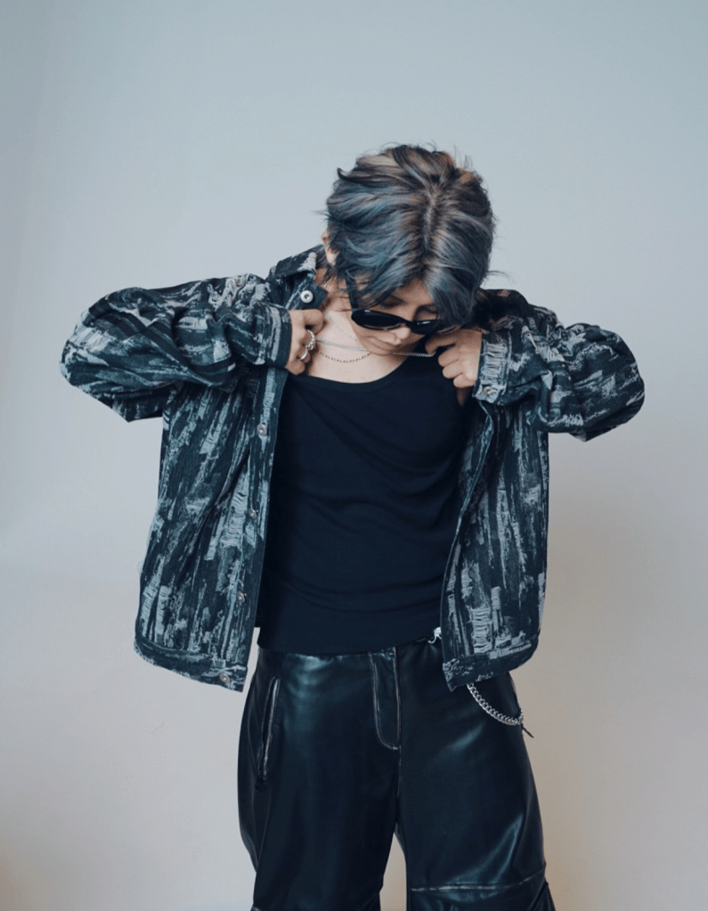 Un jeune homme avec des cheveux bleu clair, vêtu d'une veste à motifs sombres et de pantalons en cuir, ajuste sa tenue tout en regardant vers le bas.