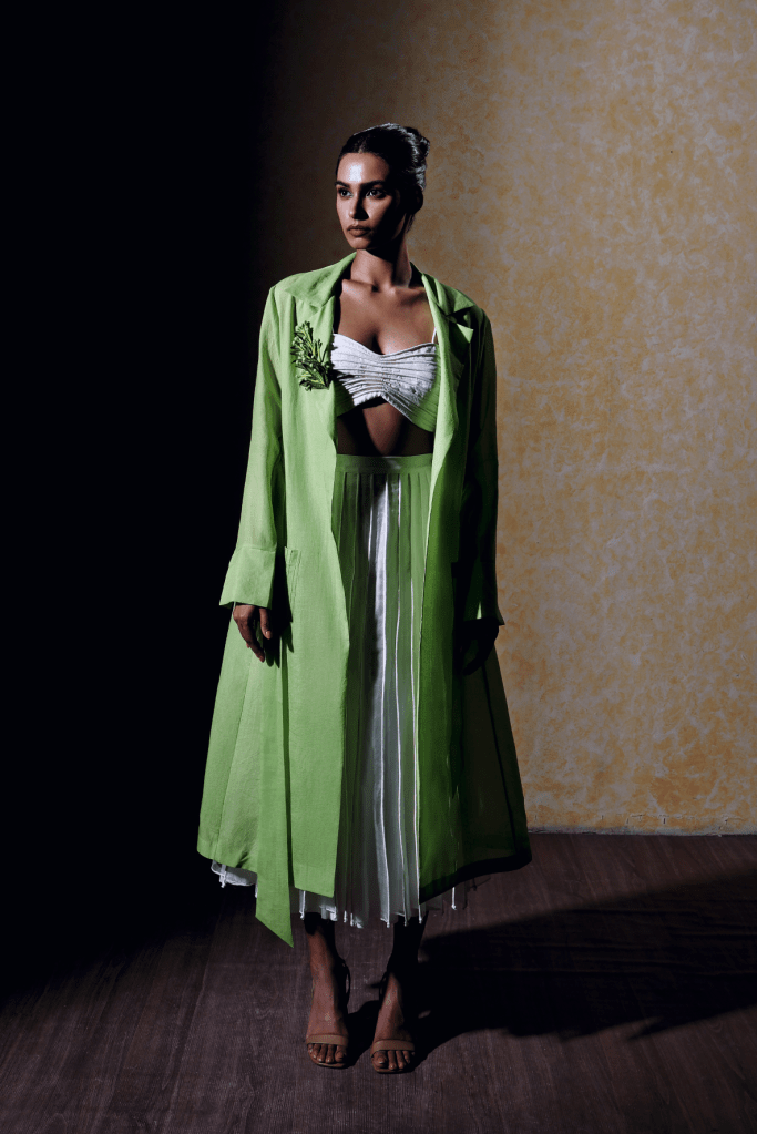 Modèle portant un manteau vert vif et une jupe fluide, avec un fond texturé et sombre.