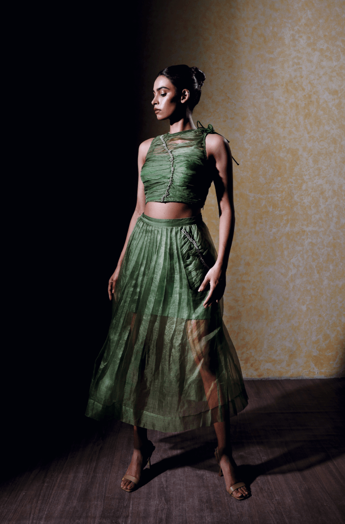 Une mannequin porte un ensemble vert constitué d'un crop top et d'une jupe longue transparente, avec un fond texturé qui met en valeur le vêtement.