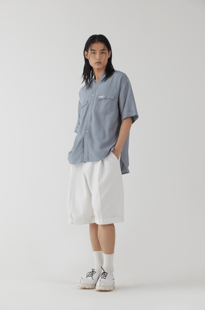 Modèle portant une chemise bleu clair et un short blanc, debout sur fond blanc, avec une expression neutre.