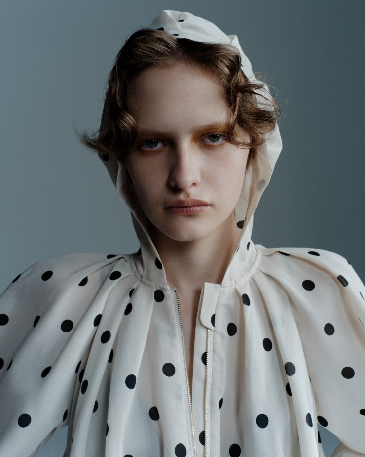 Portrait d'une femme avec un haut à pois et un capuchon, affichant une expression sérieuse sur fond neutre.