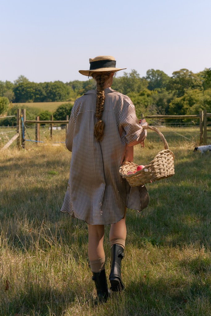 Femme de dos portant une robe à carreaux et un chapeau, tenant un panier dans un champ ensoleillé.