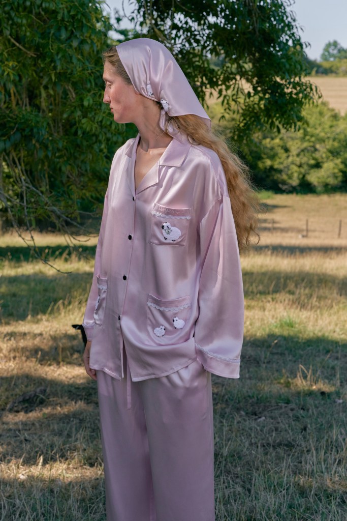 Une femme portant des vêtements de loungewear en soie rose, comprenant une chemise à poches et un foulard, se tient dans un champ avec des arbres en arrière-plan.