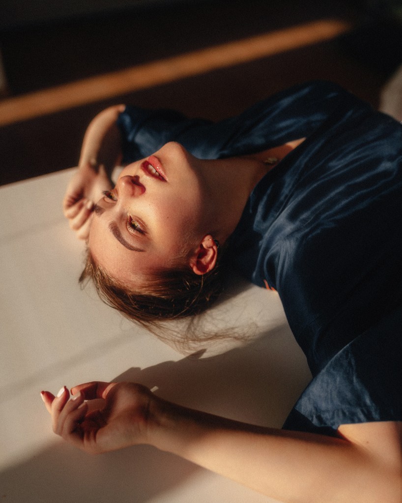 Une femme allongée sur le dos, portant une chemise bleu foncé, admirant la lumière naturelle qui éclaire son visage. Ses cheveux et ses bras sont délicatement positionnés, évoquant une ambiance tranquille et contemplative.
