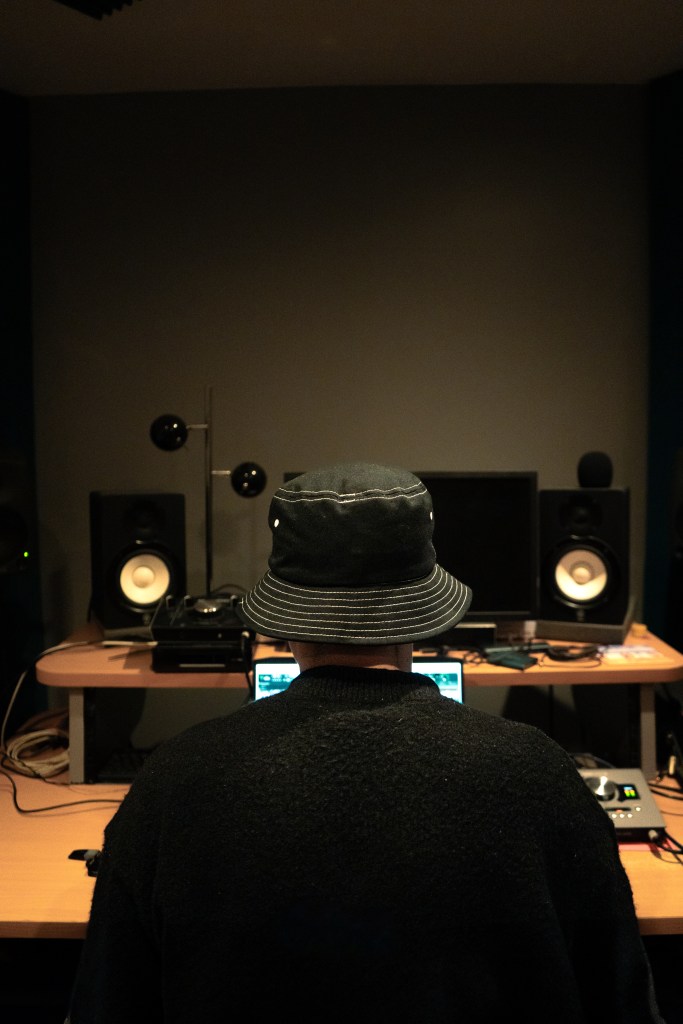 Un producteur de musique, vu de dos, concentré sur son ordinateur dans un studio d'enregistrement. Équipements audio visibles, avec des enceintes et un écran derrière lui.