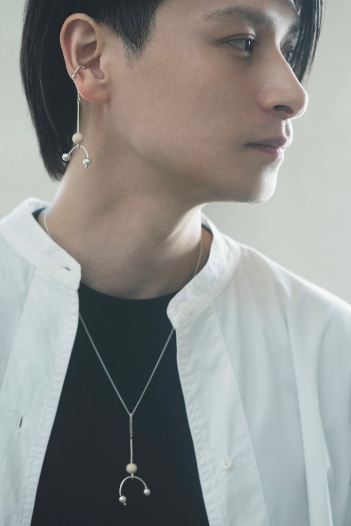 Un homme portant des boucles d'oreilles et un collier de la marque Emamima, vêtu d'une chemise blanche sur un t-shirt noir, avec un fond flou.