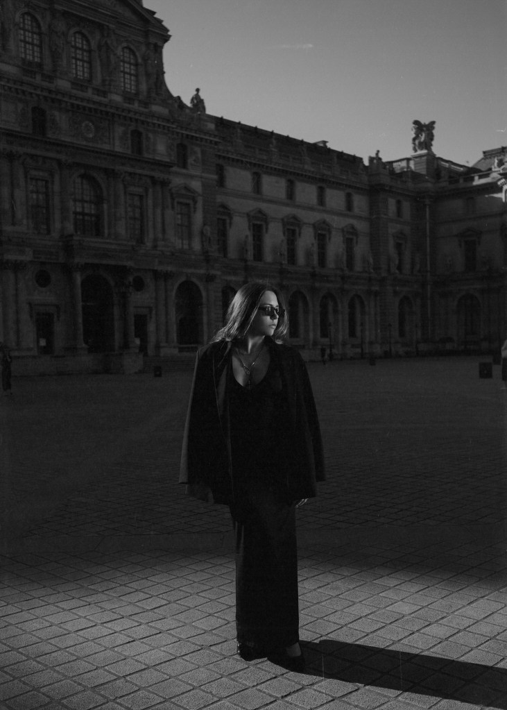 Portrait d'une femme en noir et blanc, portant des lunettes de soleil et une robe élégante, debout sur une place devant un grand bâtiment architectural à Paris.