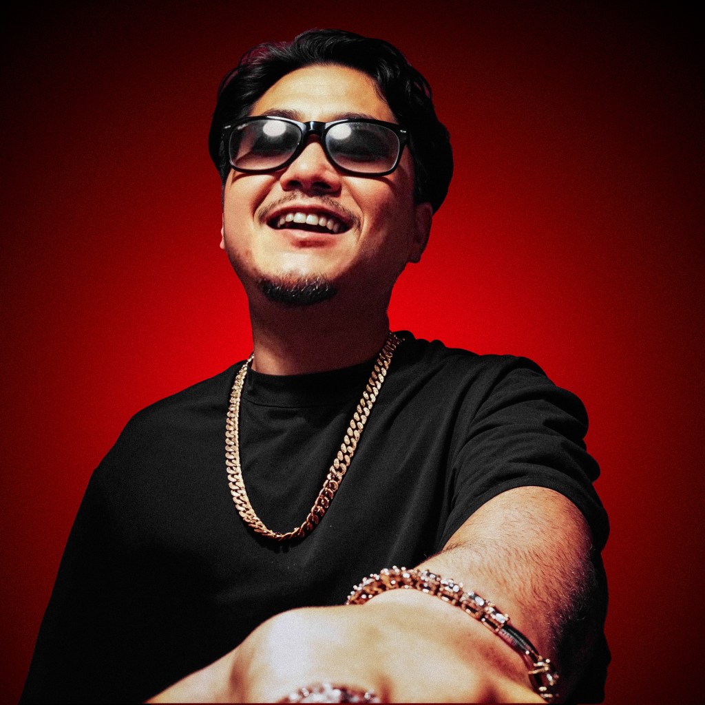 Portrait d'un homme souriant portant des lunettes de soleil et une chaîne en or, avec un fond rouge.