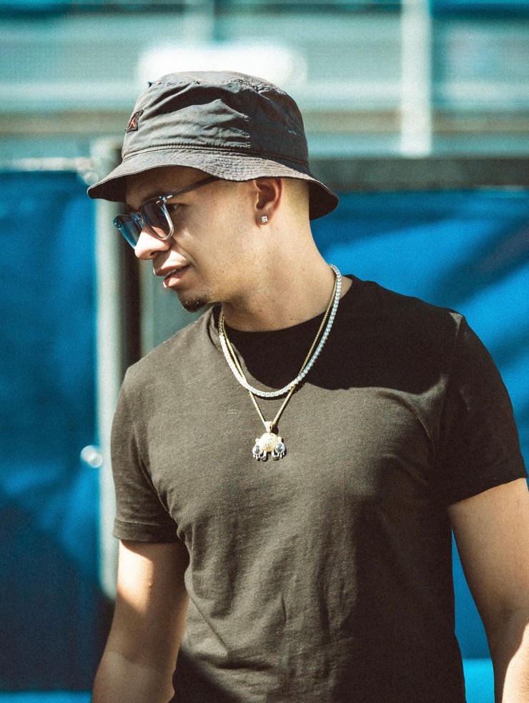 Portrait d'un homme portant un chapeau noir et des lunettes de soleil, affichant un t-shirt noir et plusieurs chaînes autour du cou, avec un fond bleu flou.