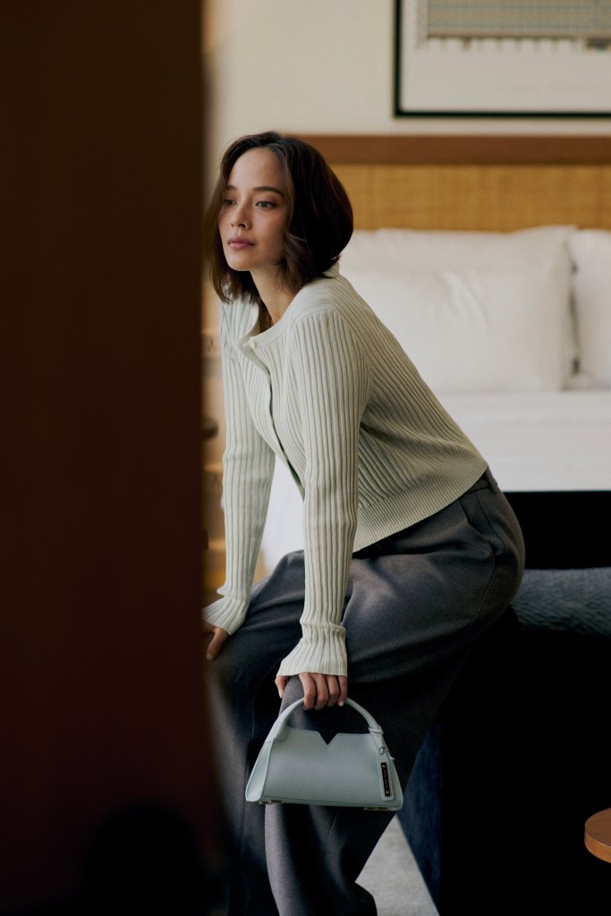 Une femme assise sur un canapé, portant un pull à manches longues de couleur claire et un pantalon gris, tenant un sac à main bleu clair inspiré par le design minimaliste.