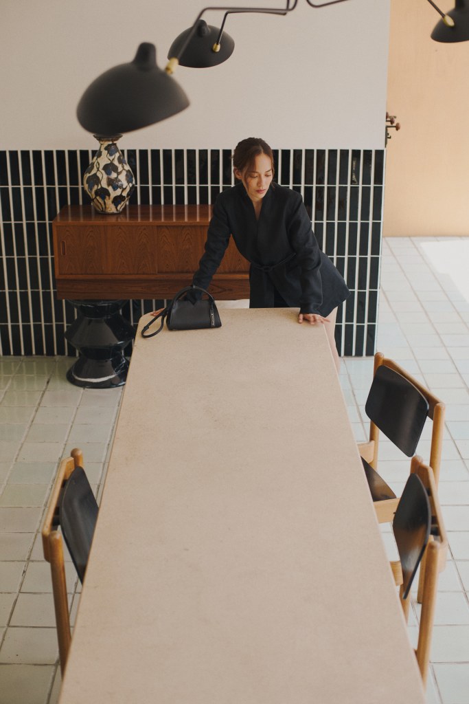 Une femme en vêtement sombre se tenant près d'une table minimaliste, avec un sac à main posé à côté d'elle, dans un intérieur moderne et épuré.