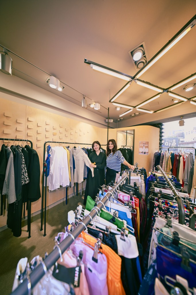 Intérieur de SHOP JINJJA à Séoul, avec deux femmes souriantes près de vêtements accrochés sur des portants. L'espace est moderne et lumineux, mettant en avant des créateurs de mode coréens.