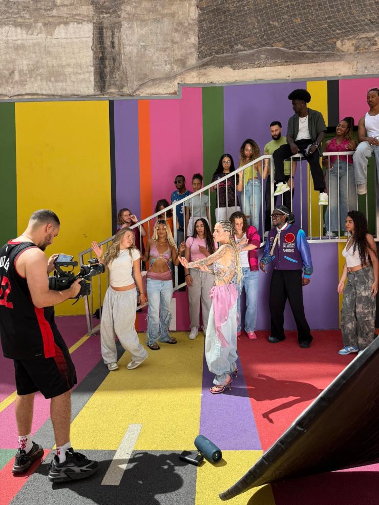 Un groupe de personnes au style éclectique se rassemble dans un décor coloré, avec une femme au centre exprimant de l'énergie et du mouvement pendant le tournage d'une vidéo musicale.