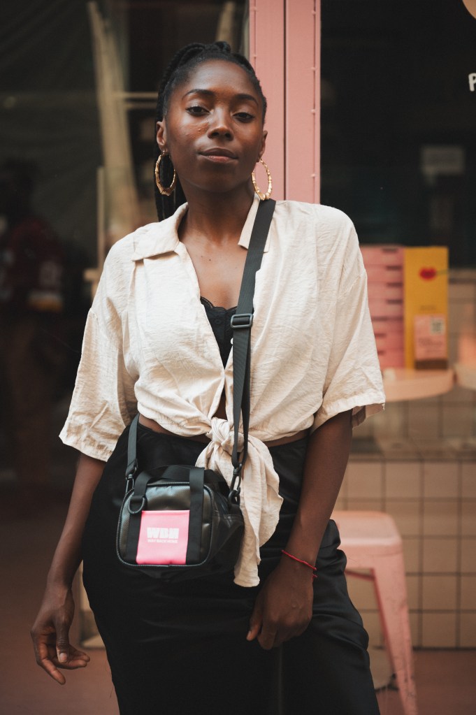Une femme élégante posant devant un mur rose, portant une chemise beige nouée et un pantalon noir, avec un sac à bandoulière de la marque Way Back Home.