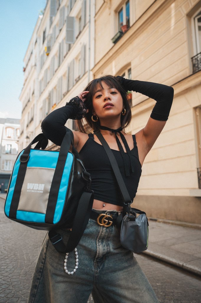 Une femme pose dans une rue parisienne, vêtue d'un haut noir et de jeans, tenant un sac de la marque Way Back Home en bleu et noir.