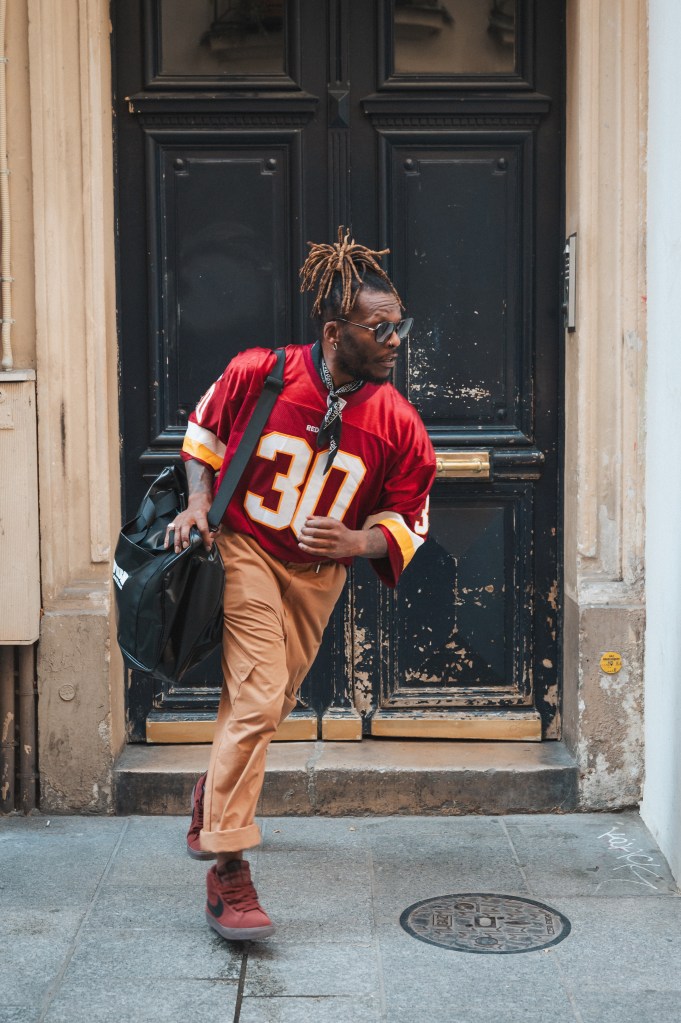 Un homme avec des dreads porte un maillot rouge, un pantalon beige et des chaussures rouges, marchant avec un sac noir devant une porte en bois sombres à Paris.