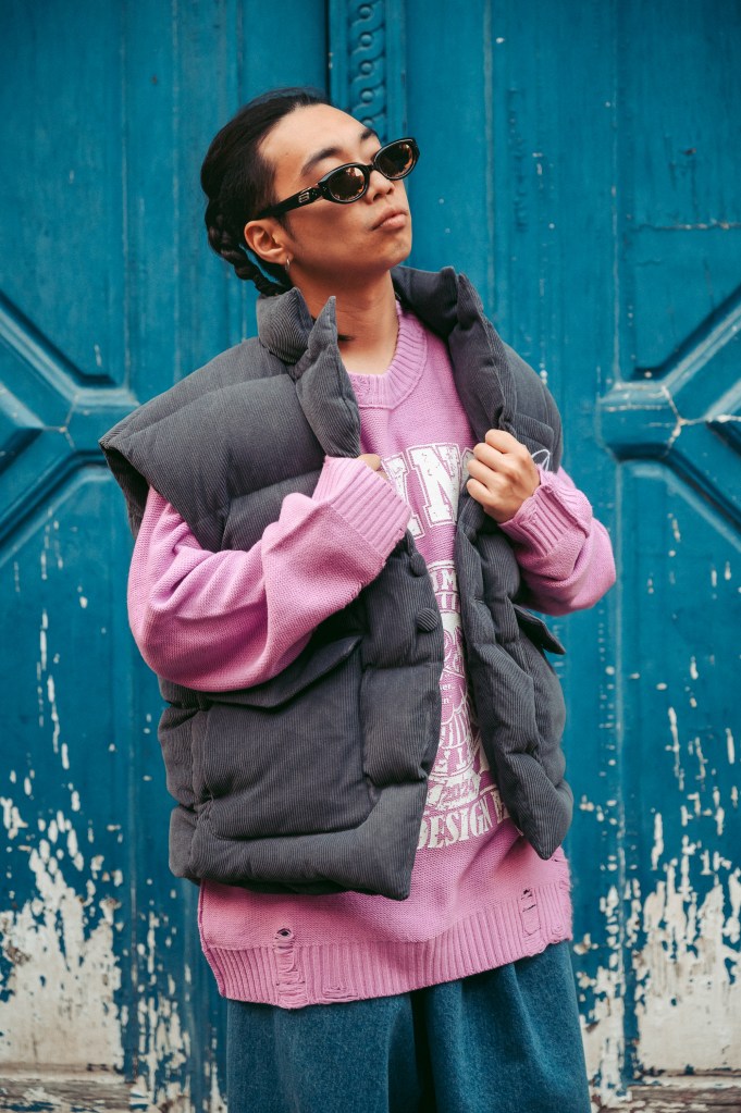 Personne portant un gilet gris et un pull rose, posant devant un mur bleu décoloré.