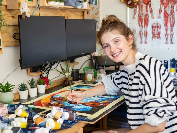 An artist smiling while painting in a creative workspace, surrounded by paint tubes, plants, and computer monitors.