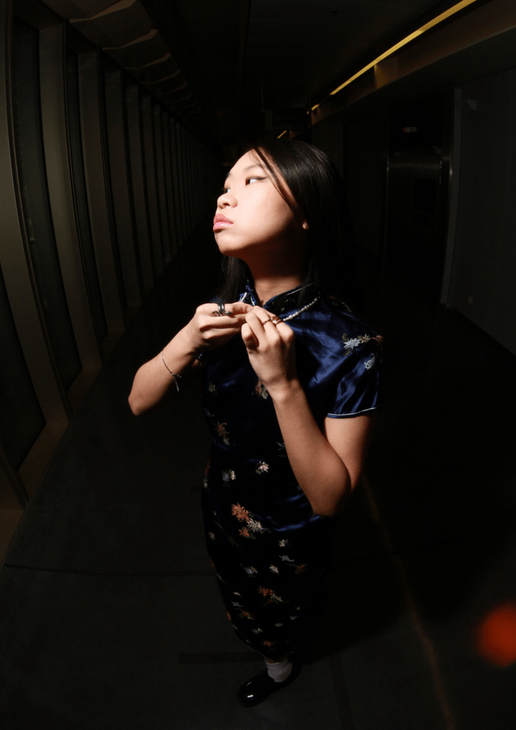 A young girl wearing a blue floral cheongsam poses confidently while adjusting her collar in a corridor with large windows.