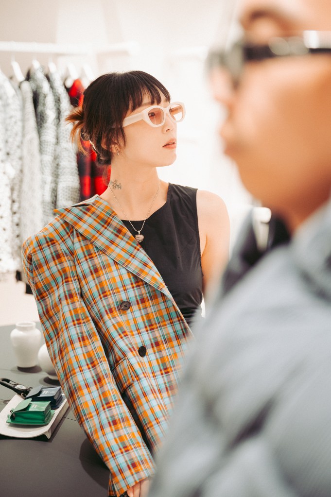 A woman with short dark hair wearing glasses and a black top is looking thoughtfully while draped in a multicolored plaid oversized blazer. A blurred figure of a person is seen in the foreground.