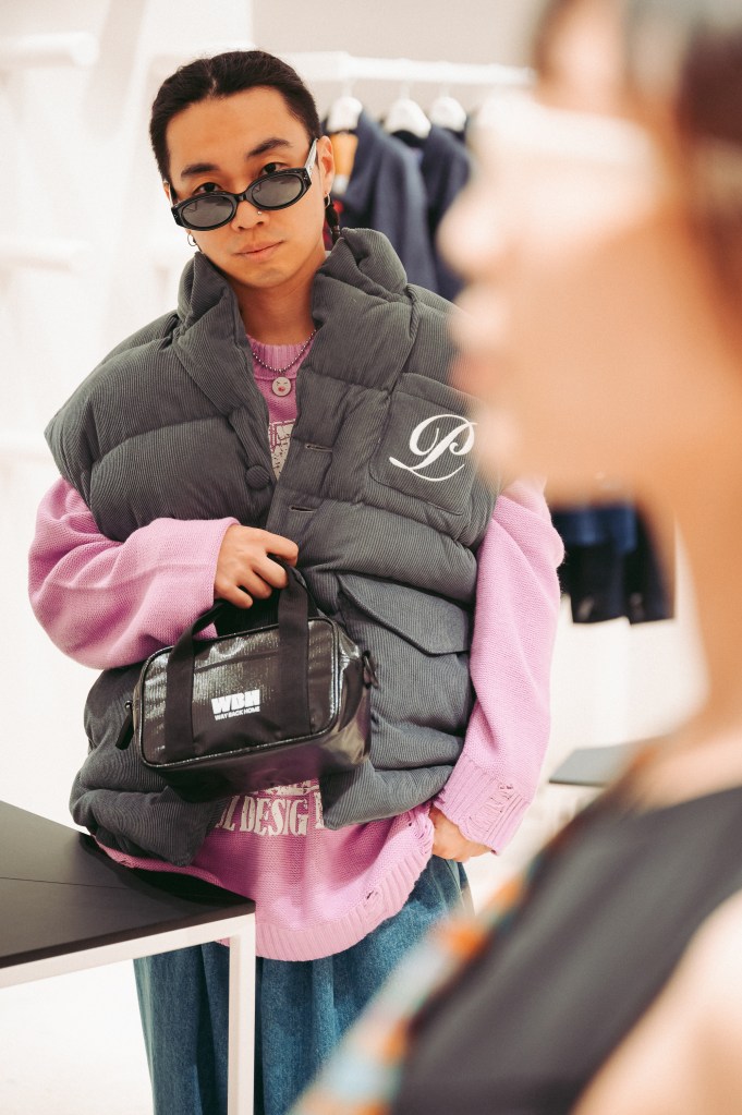 A stylish individual wearing sunglasses, a grey padded vest with a logo, and a pink sweater, holding a black bag. The background features clothing racks and a softly blurred figure, creating a fashionable atmosphere.