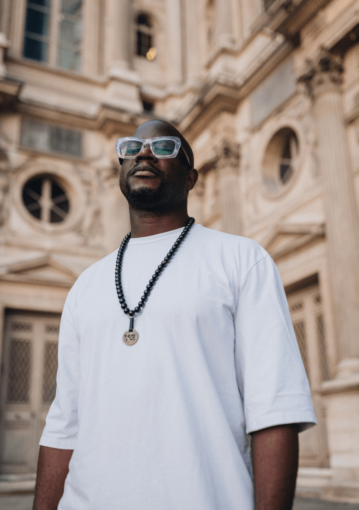 A man wearing a white t-shirt and glasses stands outdoors in front of a historic building, with a calm expression and a necklace visible around his neck.