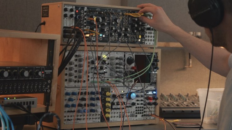A person adjusting knobs and wires on a modular synthesizer setup in a recording studio, while wearing headphones.