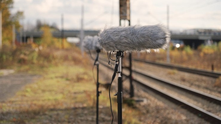 Professional audio recording equipment with furry windshields positioned along railway tracks in a natural setting.
