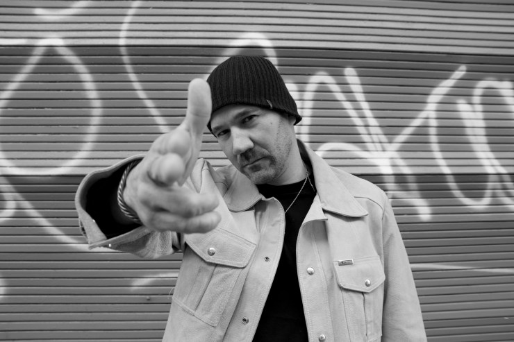 A man in a black beanie and a light-colored jacket poses against a graffiti-covered wall, extending his hand in a gesture reminiscent of pointing a finger.