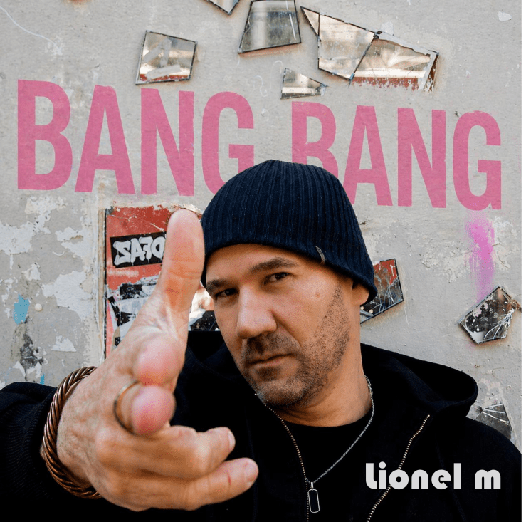 A man in a black beanie and jacket poses with his hand pointed forward, set against a wall with the words 'BANG BANG' written in pink and scattered mirror pieces.