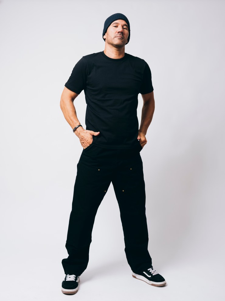 A man standing confidently with his hands in his pockets, wearing a black t-shirt, black pants, and a beanie, against a plain background.