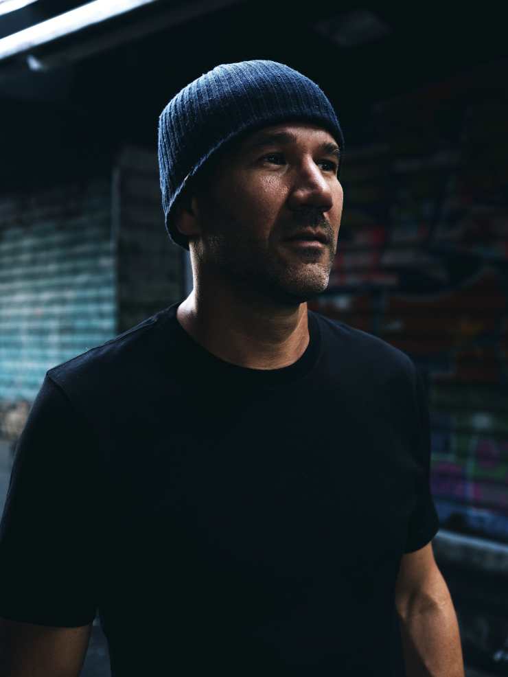 A man wearing a dark shirt and a beanie hat stands in front of a graffiti-covered wall, looking thoughtfully into the distance.