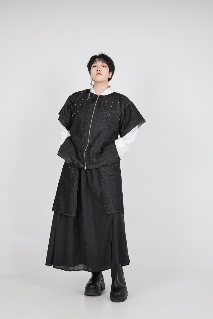 A person wearing a stylish black outfit with decorative studs, featuring a zippered top and a long skirt, standing confidently against a plain background.