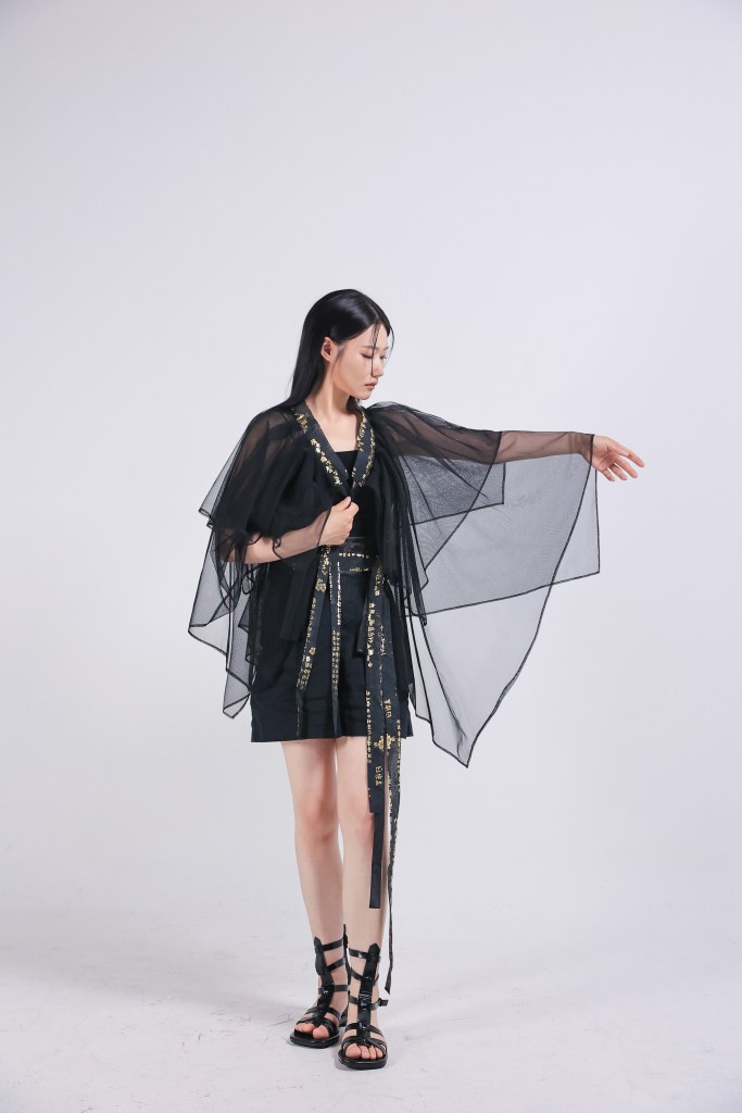A model wearing a black sheer kimono-style top over a fitted outfit, striking a dynamic pose with one arm extended.
