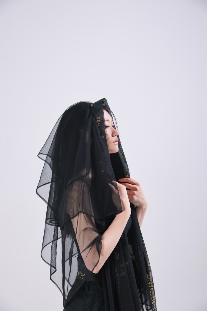 A woman wearing a black sheer veil, standing in profile with a serene expression, against a white background.