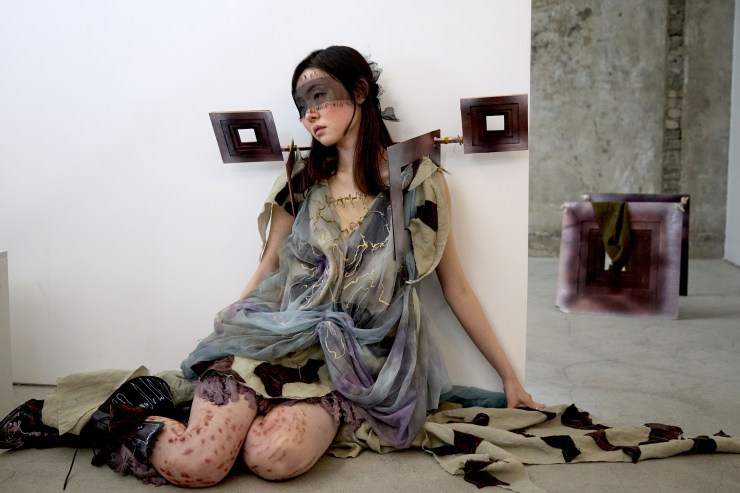 A model sitting against a white wall, wearing an artistic outfit made of layered fabric in muted tones, with intricate designs and accessories. The background features abstract sculptures in geometric shapes.