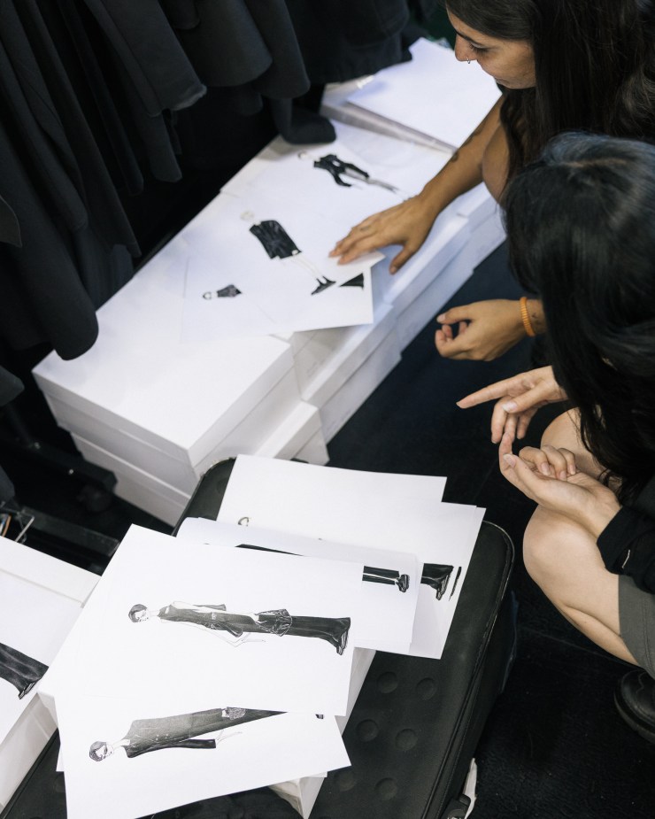 Two women examining fashion sketches of clothing designs on white paper, with black garments hanging in the background.