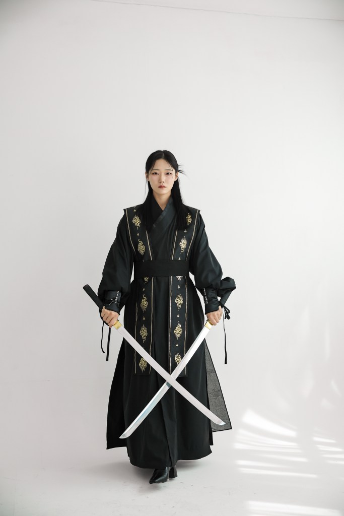 A person wearing a traditional black outfit with gold embroidery, holding two swords in a neutral pose against a white background.