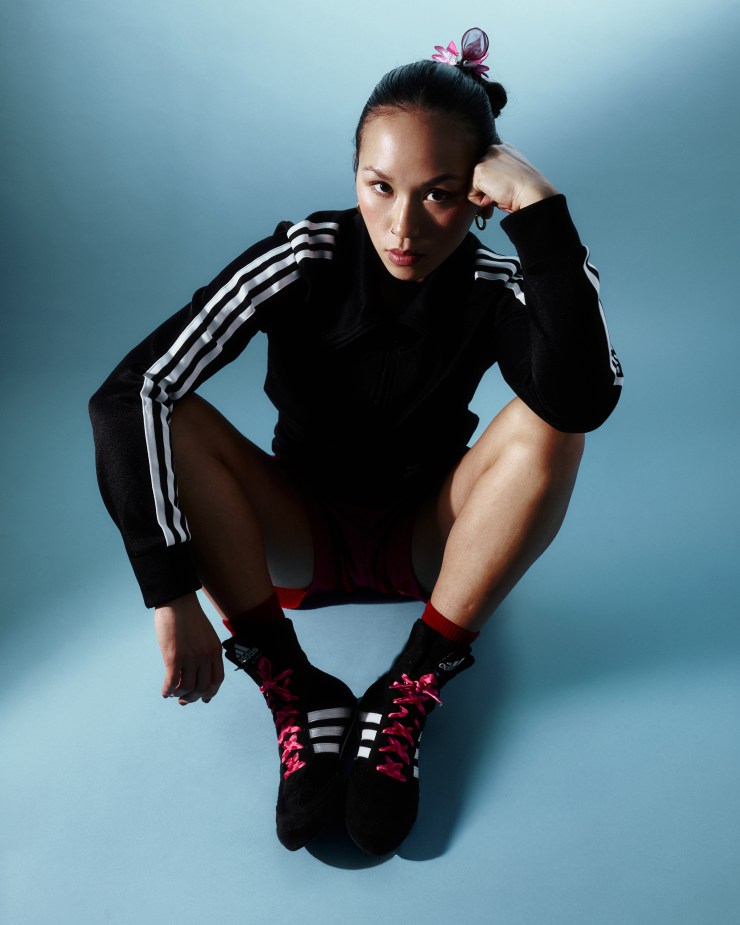 A person sitting in a squat position, wearing a black athletic jacket with three white stripes, pink athletic shorts, and black shoes with pink laces. The background is a soft blue gradient.