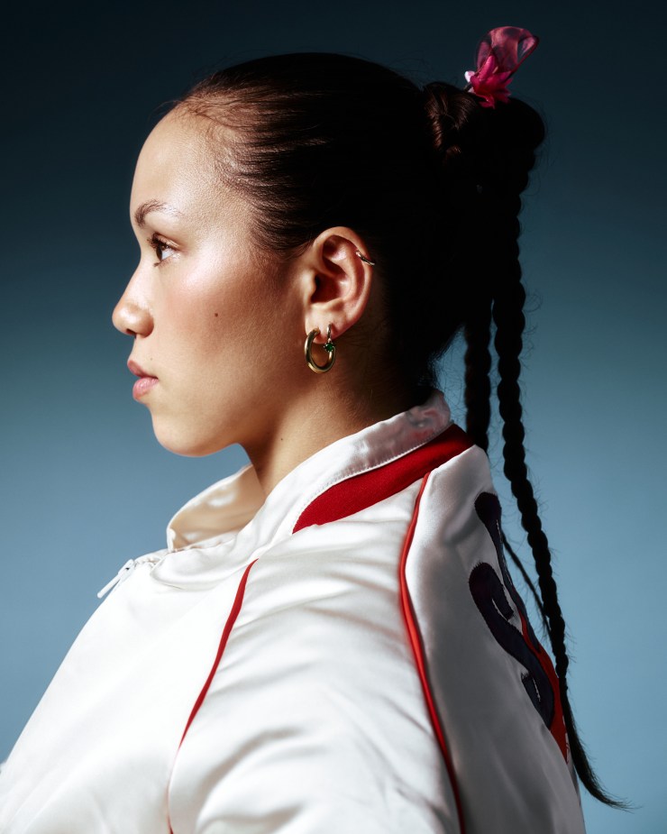 Profile view of a young woman with braided hair and a pink hair accessory, wearing a white jacket with red accents.