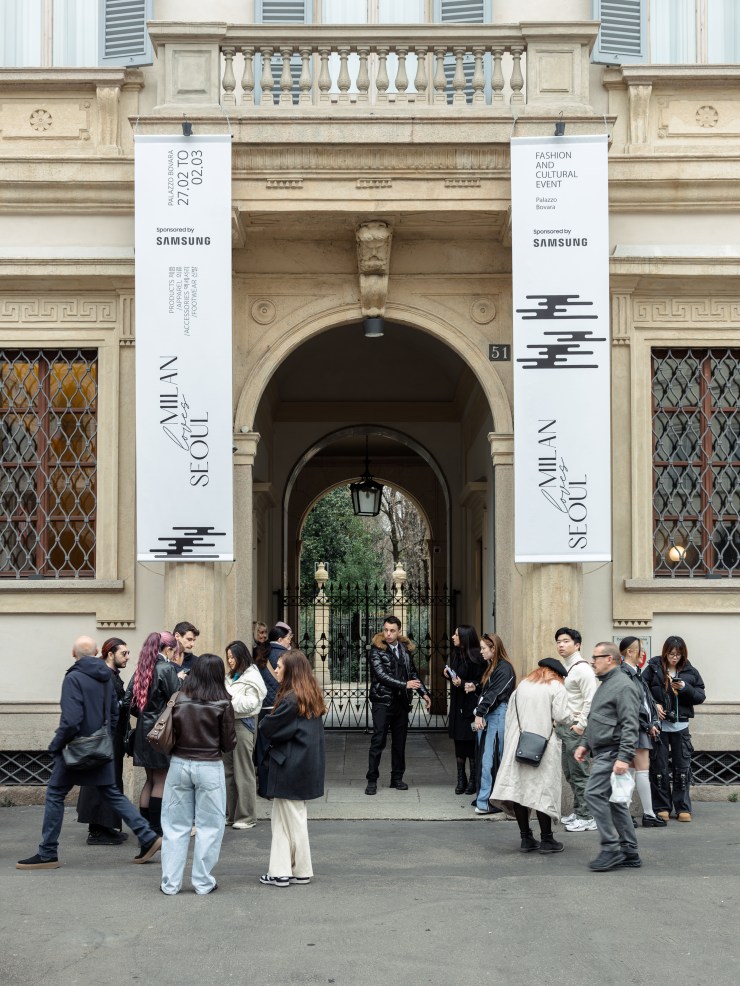 A group of people gathered in front of a historic building entrance with banners advertising a fashion and cultural event titled 'Milan Meets Seoul'. The scene captures attendees interacting as they prepare for the event.