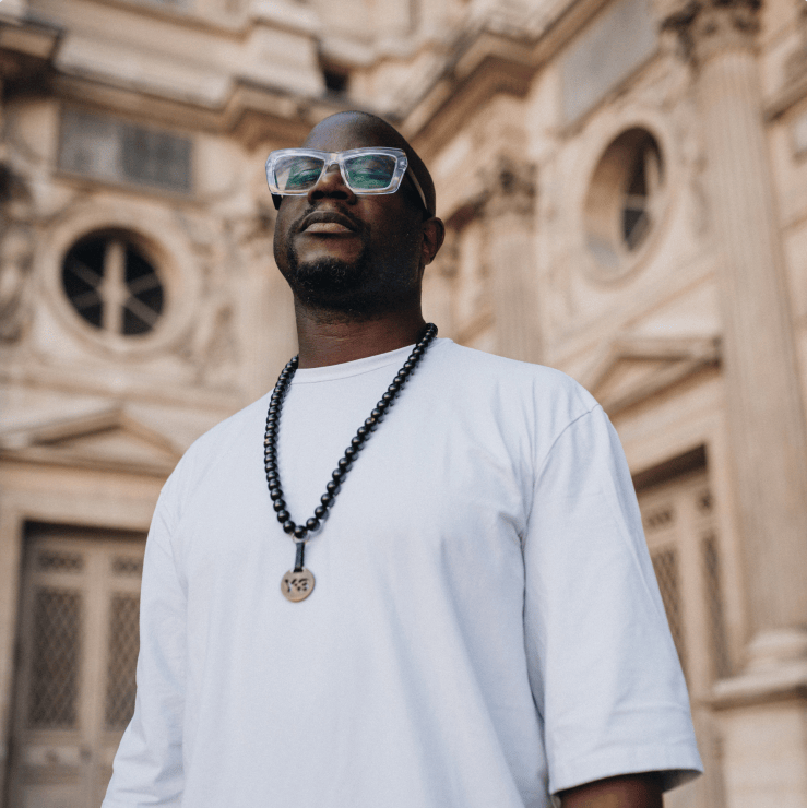 A man wearing a white t-shirt and glasses, standing outdoors in front of a historic building, with a necklace made of beads.