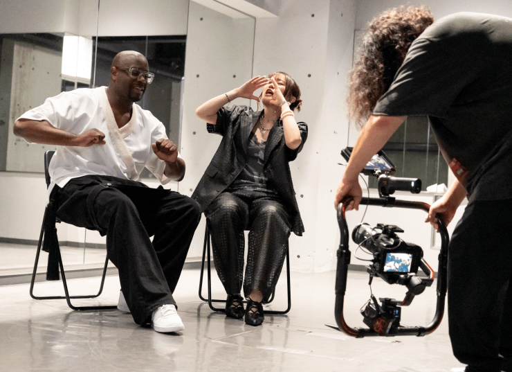 Two people sitting on chairs in a studio, one man laughing and the other woman covering her face in amusement, while a third person operates a camera.