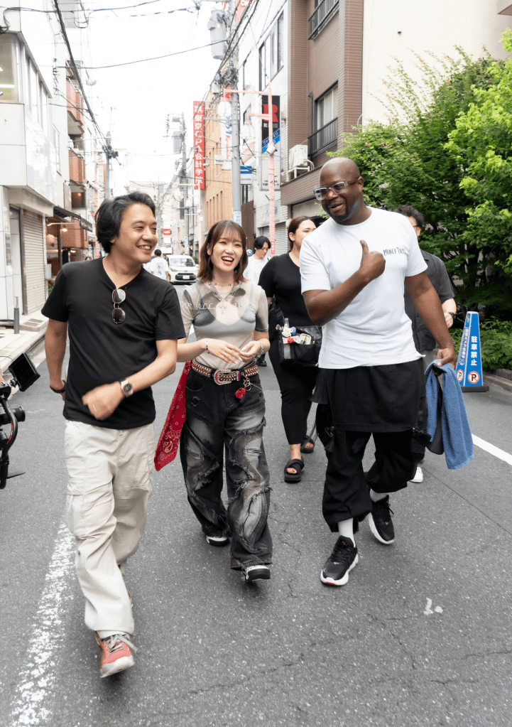 A group of four friends walking down a city street, smiling and chatting. The scene features a man in a black t-shirt and sunglasses, a woman wearing a graphic t-shirt and baggy pants with a red bandana, a second man in a white t-shirt, and another person in casual attire, surrounded by urban buildings and greenery.