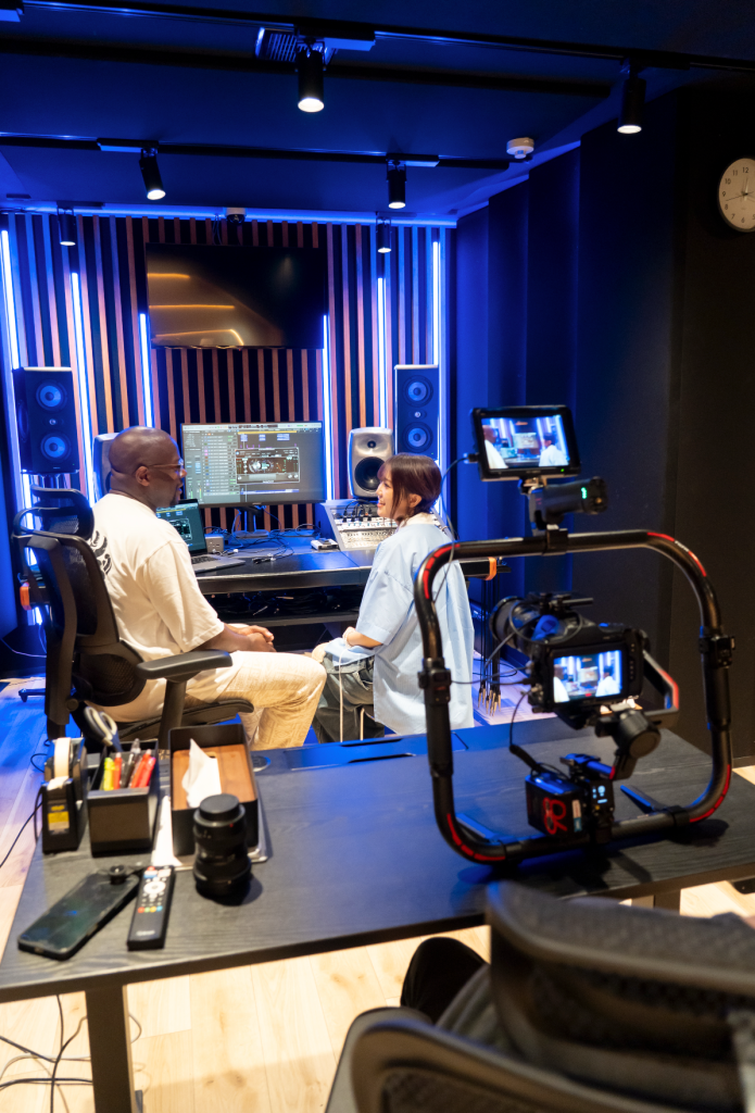 A music studio scene featuring two individuals engaged in a discussion, with a computer screen displaying audio editing software, speakers, and a camera setup. The room is illuminated with blue lighting.