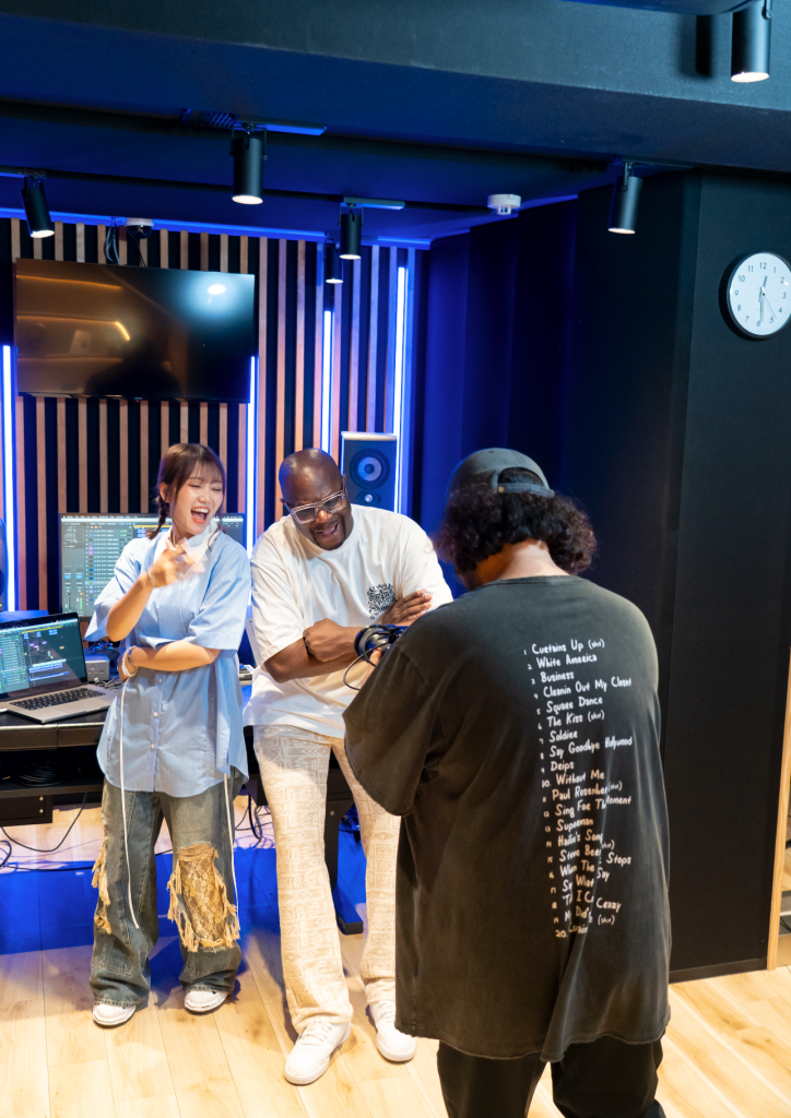 A group of three individuals in a music studio, laughing and engaging with each other. One person is holding a microphone, while a computer and audio equipment are visible in the background.