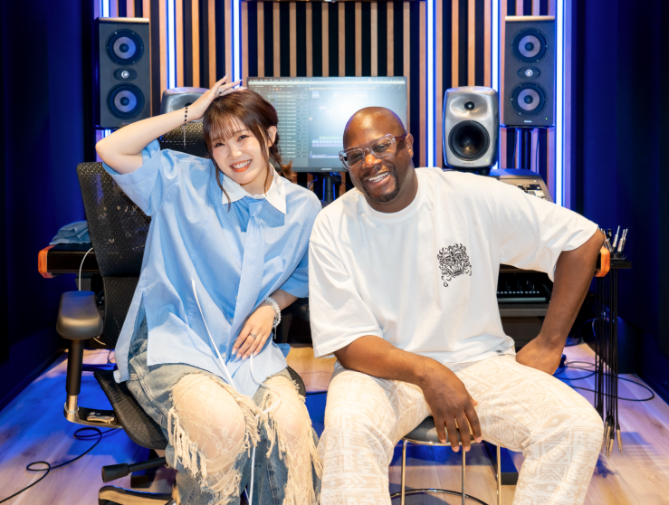 Two smiling individuals posing in a music studio, one wearing a blue shirt and the other in a white t-shirt, with recording equipment in the background.